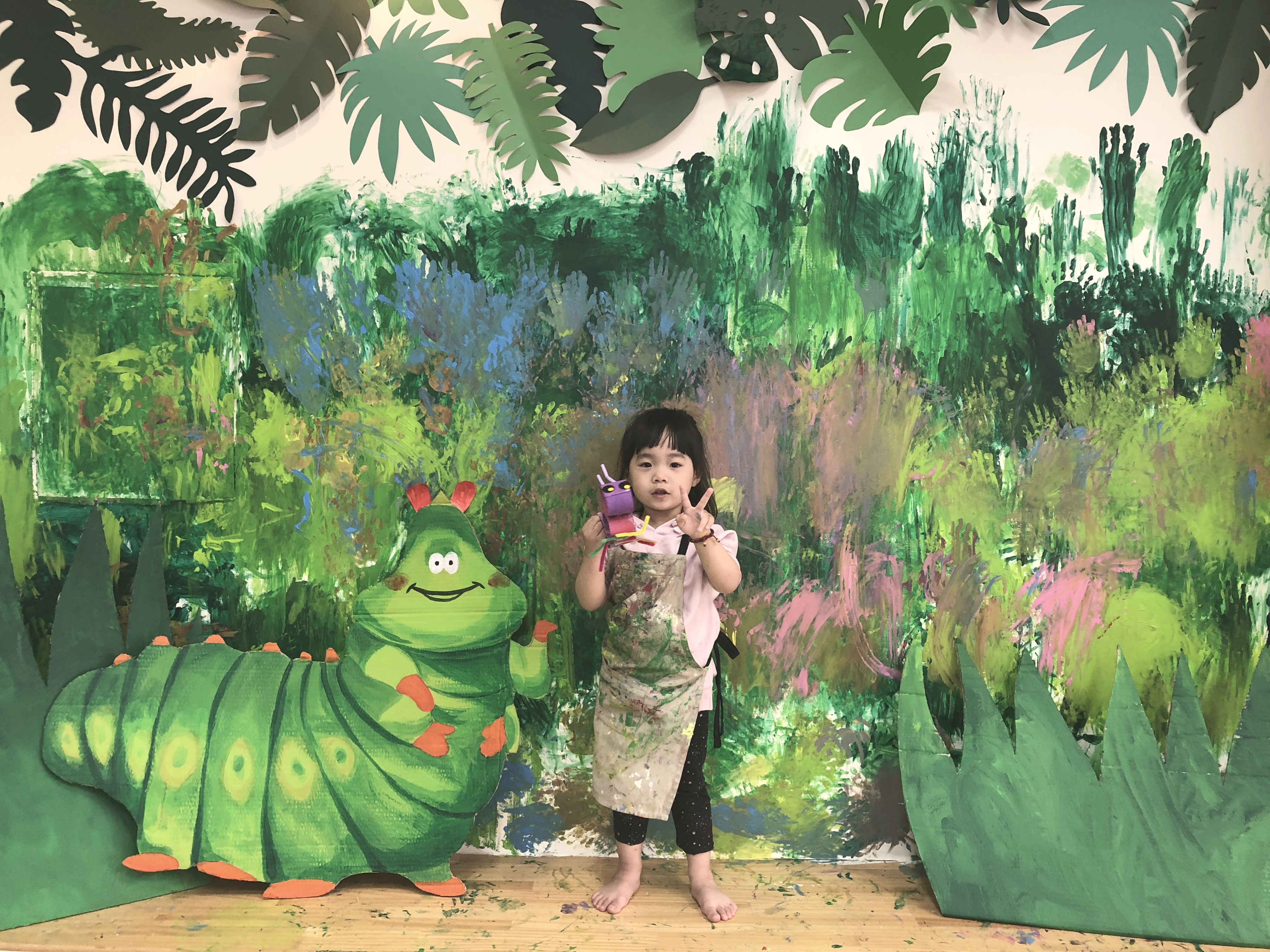 孩子站在大型森林壁畫與毛毛蟲裝置前展示作品